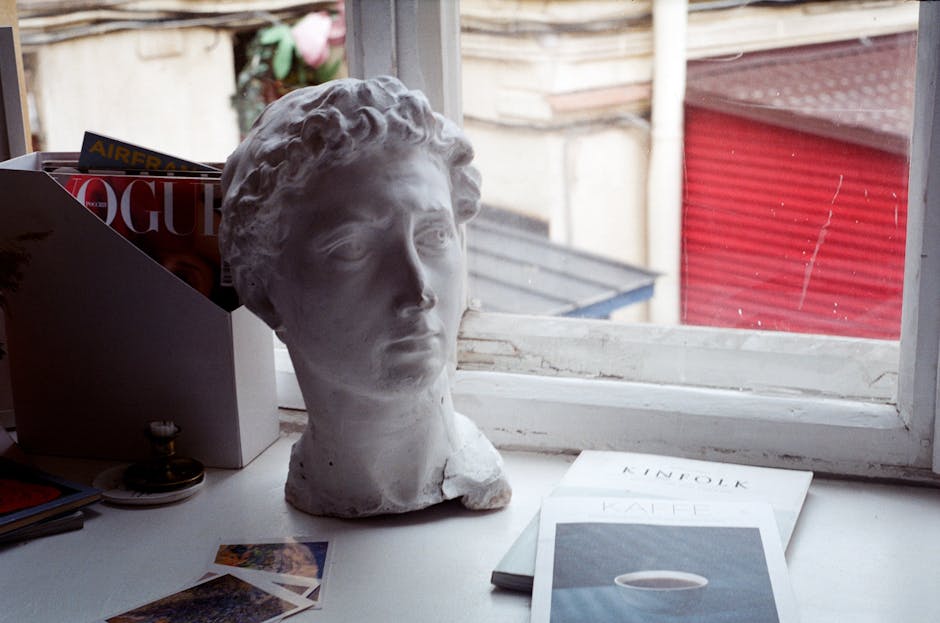 A classical bust displayed on a desk beside magazines, overlooking an urban window scene.